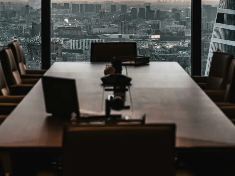 An empty boardroom table in front of a floor-to-ceiling window looking over a city at dusk.